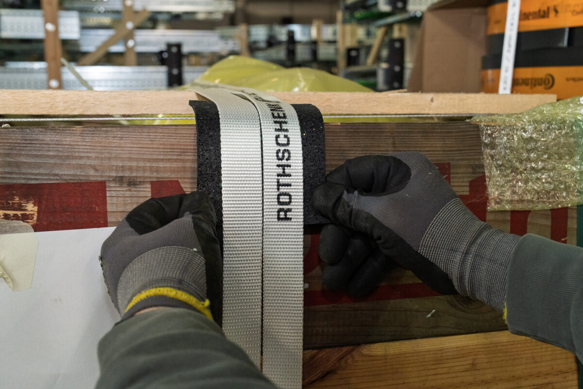 Nahaufnahme von behandschuhten Händen, die einen Sicherungsgurt über einem Kantenschutz an einer Holzkiste festziehen. Fachgerechte Ladungssicherung durch das mobile Verpackungsteam der MHS GmbH.