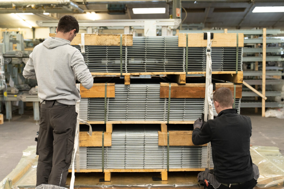 Zwei Mitarbeiter der MHS GmbH sichern schwere Gitterroste auf spezialgefertigten Transportpaletten mit Spanngurten für den sicheren Export.