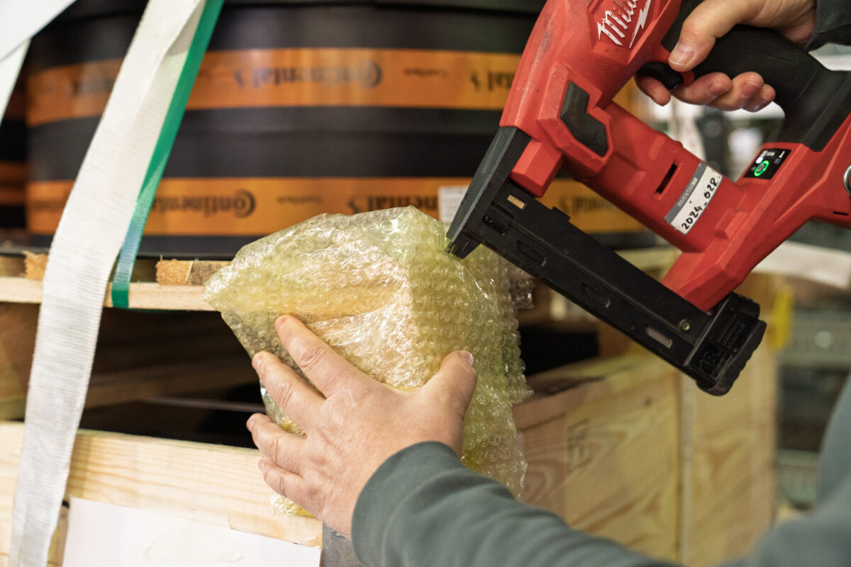 Nahaufnahme: Ein Mitarbeiter der MHS GmbH befestigt mit einem roten Akku-Nagler Polstermaterial an einer Holzkiste. Fachgerechte Verpackung direkt beim Kunden vor Ort.